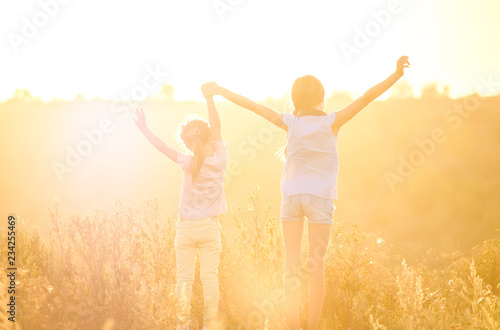 Little smiling girls stand on sunshine evening field with joyfully raised hands