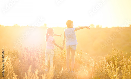 Little girls stand by holding hands looking on sunshine evening field pointing to right