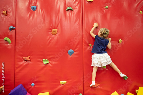 Little blond girl climbing by the wall in the play room among colorful soft cubes