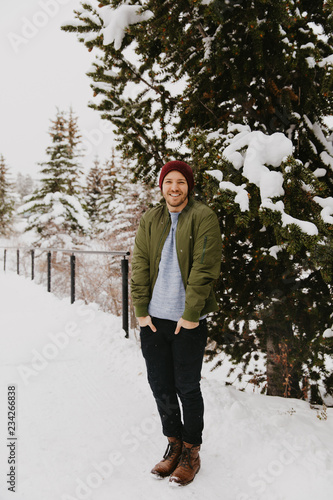 Young Trendy Man in Green Bomber Jacket Enjoying the Winter Snow on a Small Bridge in Colorado © MeganMahoneyPhotos