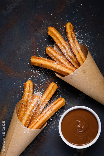 Traditional churros sticks in paper bag with sugar powder cinnamon and bowl of chocolate dip on dark table top view photo