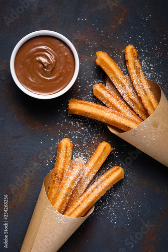 Fried traditional churro sticks with sugar powder cinnamon and bowl of chocolate dip on dark table top view photo