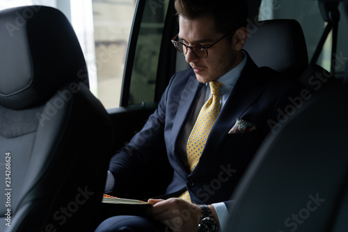 Young businessman sitting on back seat of the car, while his chauffeur is driving automobile.