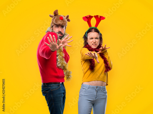 Couple dressed up for the christmas holidays is a little bit nervous and scared stretching hands to the front on yellow background