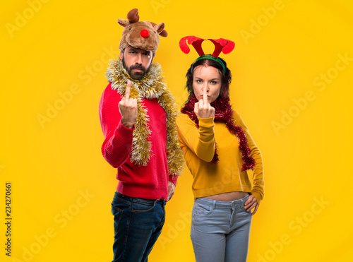 Couple dressed up for the christmas holidays making horn gesture. Negative expression on yellow background
