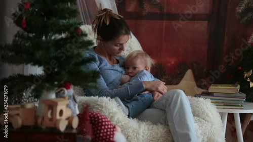 Mother breastfeeding her toddler son sitting in cozy armchair near Christmas tree, wintertime