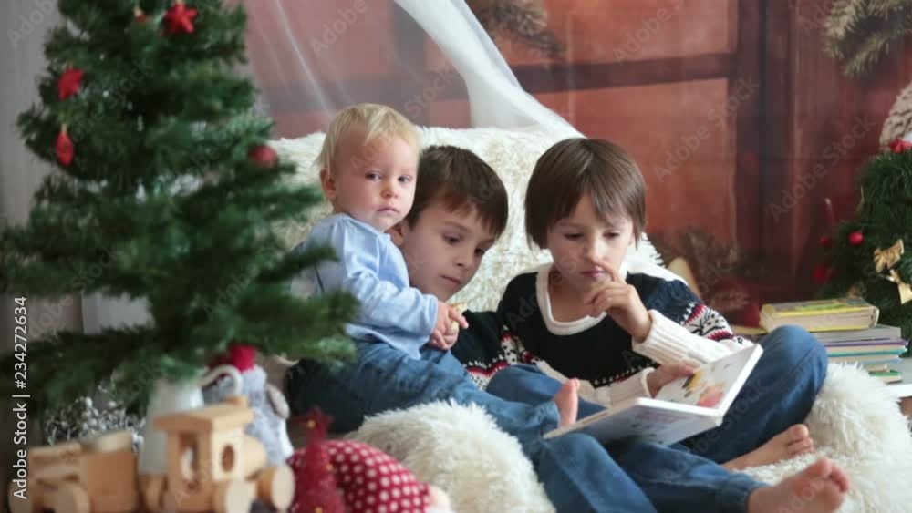 Three children, boy brothers, sitting in rocking chair in cozy living room with christmas decoration, reading a book, smiling Three children, boy brothers, sitting in rocking chair in cozy living room with christmas decoration, reading a book, smiling