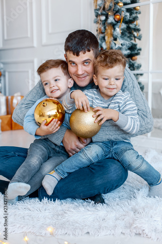 A a daddy with his two sons twins in front of the christmas tree  with candles and gifts hugging and having fun. New year's eve. Cozy holiday at the fir-tree. love, happiness and big family concept