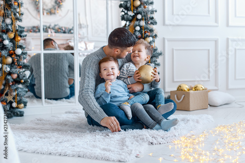 A a daddy with his two sons twins in front of the christmas tree  with candles and gifts hugging and having fun. New year's eve. Cozy holiday at the fir-tree. love, happiness and big family concept