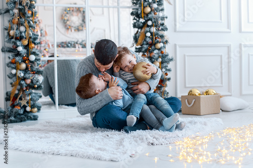 A a daddy with his two sons twins in front of the christmas tree  with candles and gifts hugging and having fun. New year's eve. Cozy holiday at the fir-tree. love, happiness and big family concept