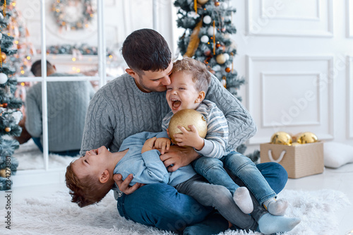 A a daddy with his two sons twins in front of the christmas tree  with candles and gifts hugging and having fun. New year's eve. Cozy holiday at the fir-tree. love, happiness and big family concept
