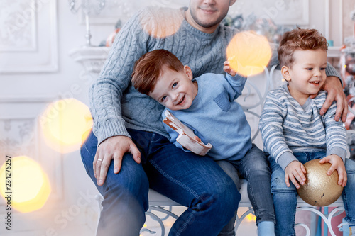 A a daddy with his two sons twins in front of the christmas tree  with candles and gifts hugging and having fun. New year's eve. Cozy holiday at the fir-tree. love, happiness and big family concept