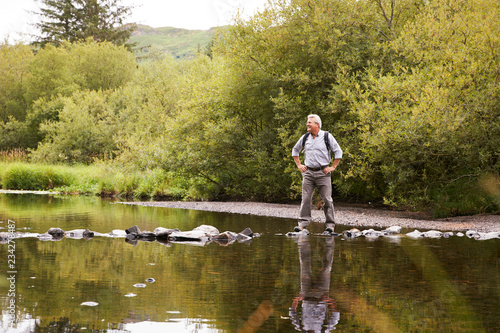 Senior Man Crossing River Whilst Hiking In UK Lake District