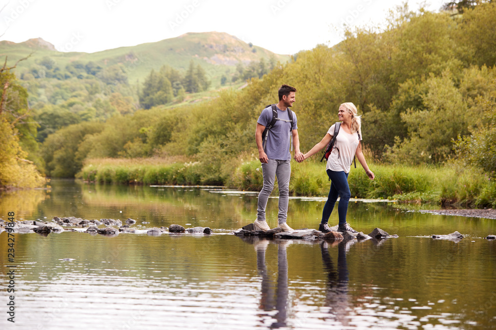 Couple Crossing River Whilst Hiking In UK Lake District