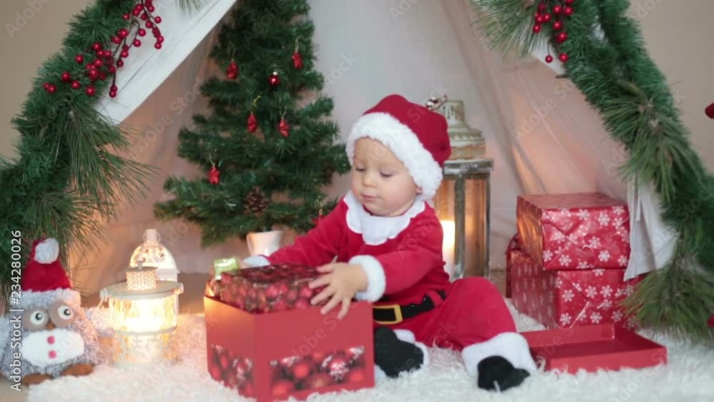 Adorable little toddler baby boy dressed in canta claus costume, playing at home in front of teepee decorated for Christmas Adorable little toddler baby boy dressed in canta claus costume, playing at home in front of teepee decorated for Christmas
