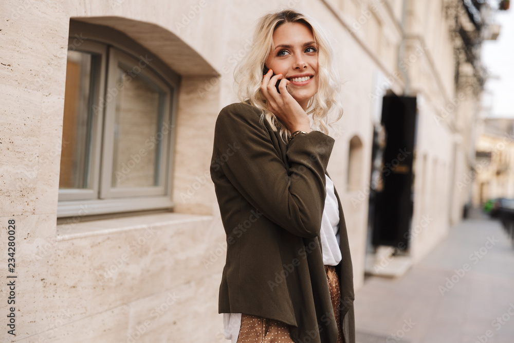 Fototapeta premium Image of blond woman wearing jacket using mobile phone for call, while walking through city street