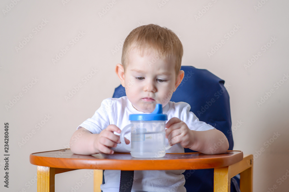 Baby boy drinking water studying a plastic bottle while sitting on a chair in the kitchen. Baby boy drinking water studying a plastic bottle while sitting on a chair in the kitchen.