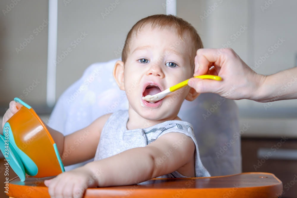 Little baby boy eats opening his mouth wide sitting on a chair in the kitchen. Mom feeds holding a spoon of porridge