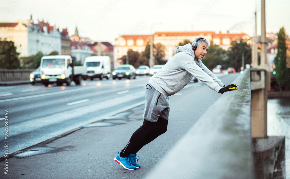 Mature male runner with headphones stretching outdoors on the bridge in Prague city.