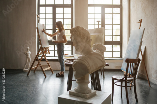 Artist at the working process. Young woman creating the painting. Workshop room with light and classical sculpture busts