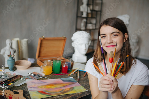Adorable young artist girl holding brushes and smiling. Creative workshop room at the background