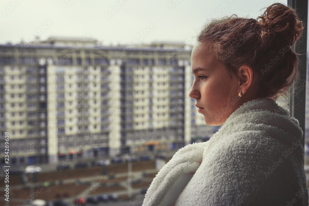 young girl looks out the window into the city, pensive, sad, turning into a warm cozy white blanket. With copy space. young girl looks out the window into the city, pensive, sad, turning into a warm cozy white blanket. With copy space.