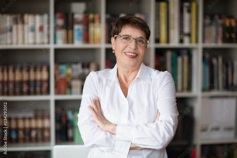 Portrait of senior businesswoman wearing glasses head shot in a white shirt, crossed hands looking at the camera with a warm friendly smile against the background of a bookcase Portrait of senior businesswoman wearing glasses head shot in a white shirt, crossed hands looking at the camera with a warm friendly smile against the background of a bookcase