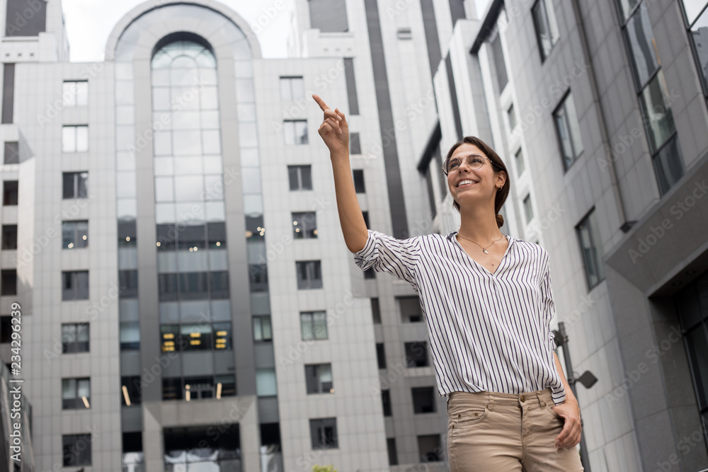 Smiling european business woman pointing up and looking away over gray city street business center background. Offise worker showing the way, official style. Look here! Over there. This is my choise