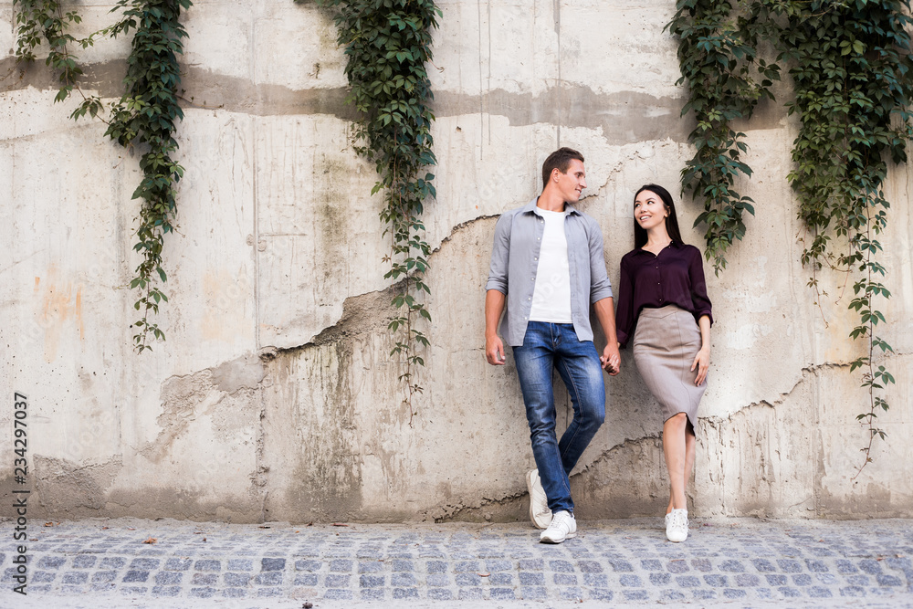 Male and female hipsters walking on grey wall background, students looking to each other and smiling, fll in love concept. Street lifestyle, multiethnic couple, asian woman and aucasian man