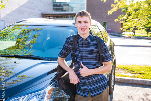 Happy male teenager leaning against his car in a high school parking lot.
