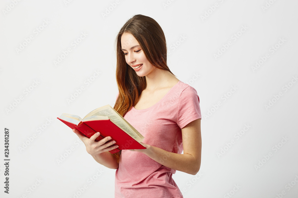 Beautiful long haired girl dressed in a pink t-shirt reads a book on a white background in the studio Beautiful long haired girl dressed in a pink t-shirt reads a book on a white background in the studio
