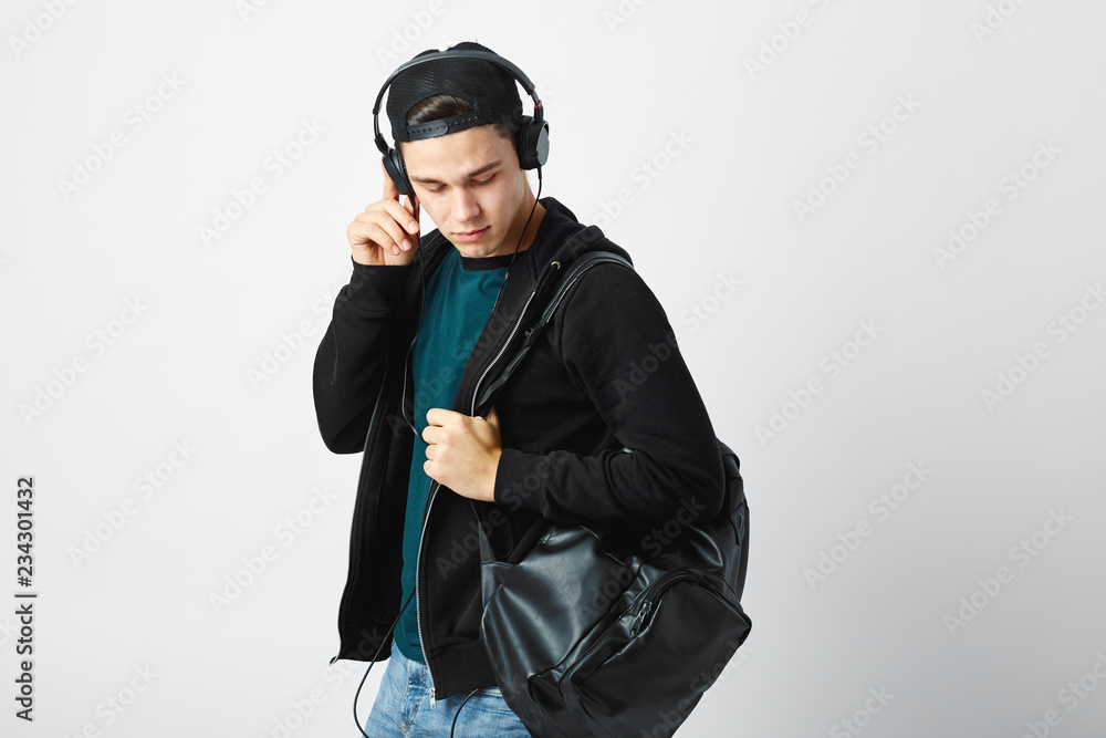 Guy with a black backpack on his shoulder and headphones on his head dressed in a dark t-shirt, jeans, sweatshirt and a cap uses mobile phone in the studio on a white background Guy with a black backpack on his shoulder and headphones on his head dressed in a dark t-shirt, jeans, sweatshirt and a cap uses mobile phone in the studio on a white background