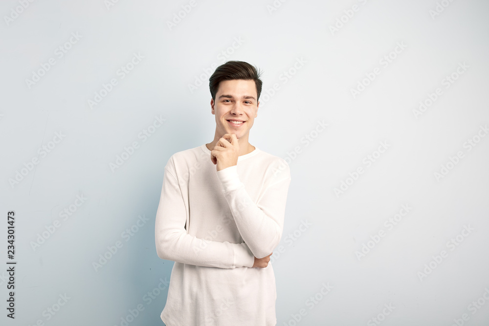 Smiling dark-haired guy dressed in a white long sleeve t-shirt and jeans stands and keeps his hand on his chin on a white background Smiling dark-haired guy dressed in a white long sleeve t-shirt and jeans stands and keeps his hand on his chin on a white background