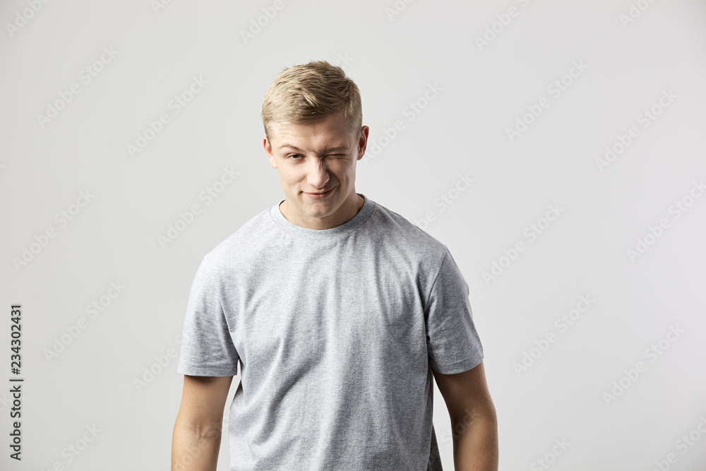 Winking blond guy dressed in a white t-shirt stands on the white background in the studio Winking blond guy dressed in a white t-shirt stands on the white background in the studio