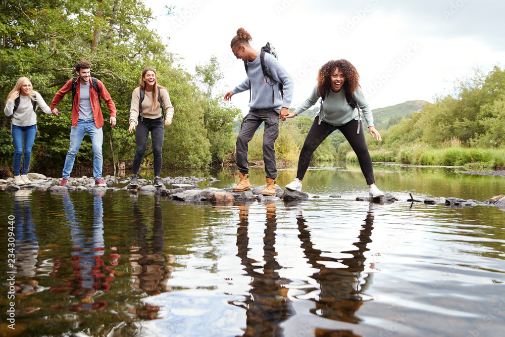 Multi ethnic group of five young adult friends laughing as they balance on rocks to cross a stream during a hike Multi ethnic group of five young adult friends laughing as they balance on rocks to cross a stream during a hike