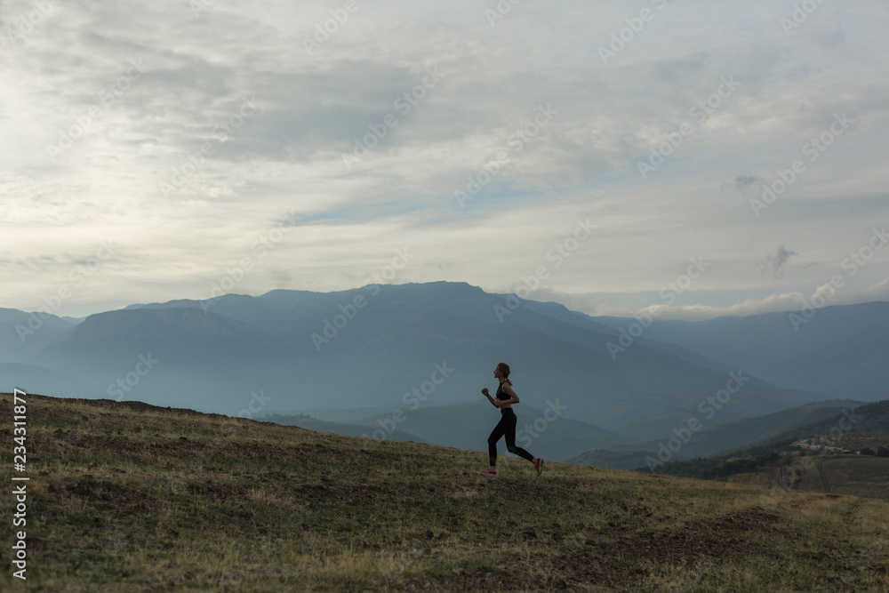 Slim girl wearing black sport outfit is jogging in mountains, beautiful landscape, cloudy sky, foggy hills.