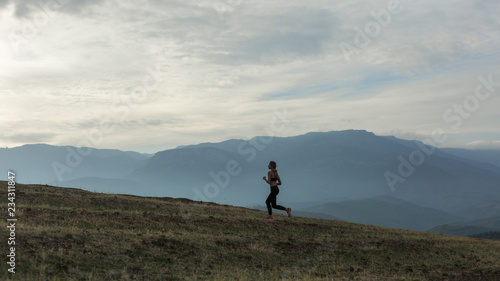 Slim girl wearing black sport outfit is jogging in mountains, beautiful landscape, cloudy sky, foggy hills.