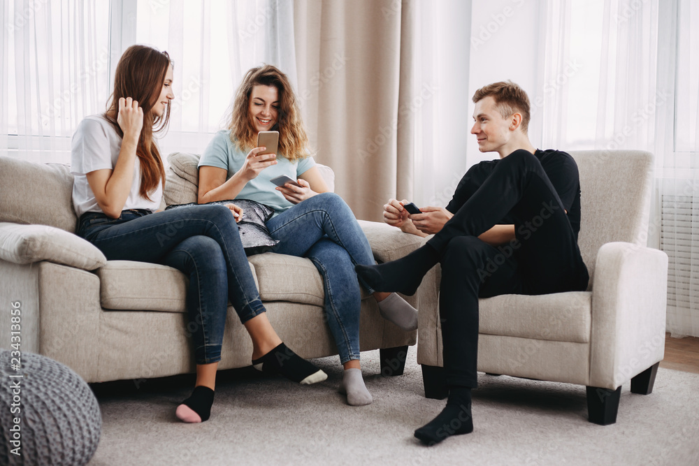 young happy friends laughing using mobile phone app together sitting in living room. leisure, social media, technology, fun concept. young happy friends laughing using mobile phone app together sitting in living room. leisure, social media, technology, fun concept.