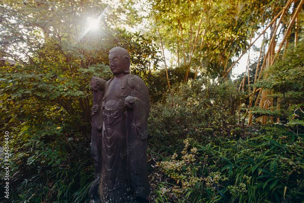A weathered, life size stone statue of a Buddhist monk holding a walking staff and wearing prayer beads meditates under green trees in japanese garden. Relax and mind calm concept. Copy space.