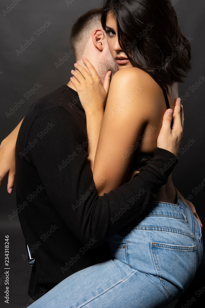Beautiful couple sitting on a white cube. Man passionately hugs and kisses in neck a girl wearing jeans and bra. Casual style. Lifestyle, love, fashion, commercial design Beautiful couple sitting on a white cube. Man passionately hugs and kisses in neck a girl wearing jeans and bra. Casual style. Lifestyle, love, fashion, commercial design