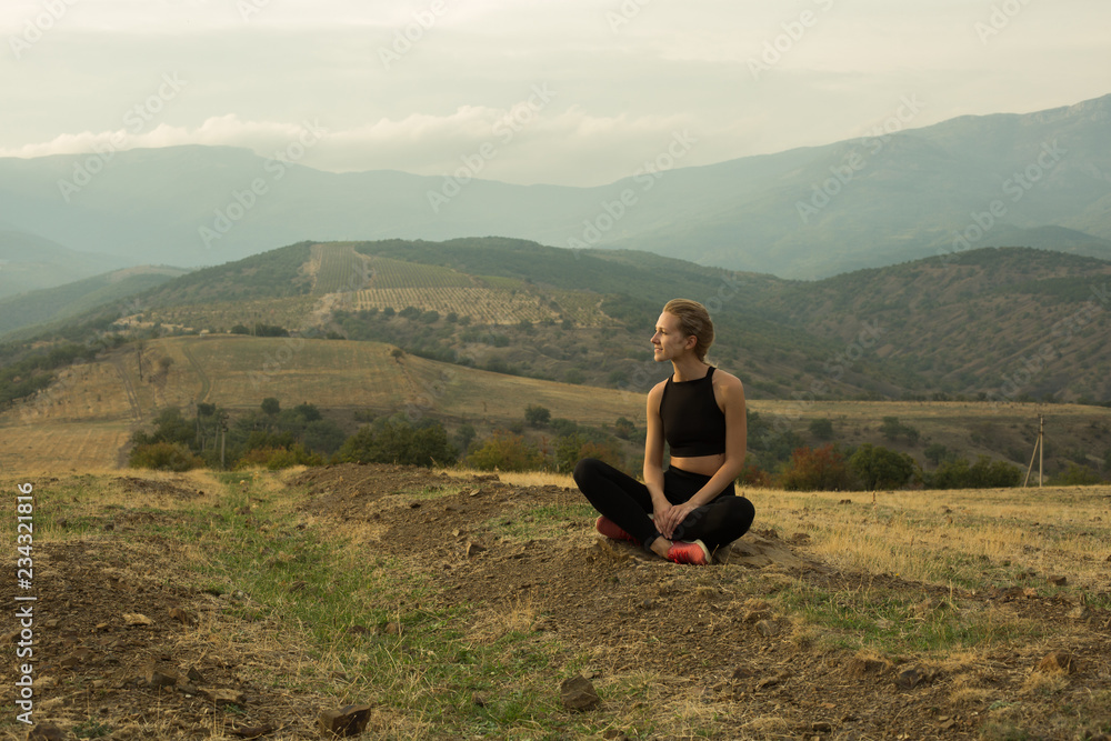 Naklejka premium Smiling young girl is squatting crosslegged among dried yellow grass, meadow, in mountains, cloudy sky