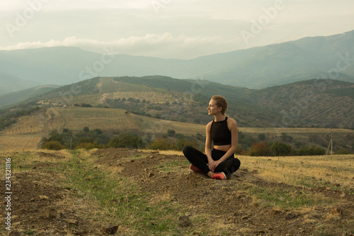 Smiling young girl is squatting crosslegged among dried yellow grass, meadow, in mountains, cloudy sky