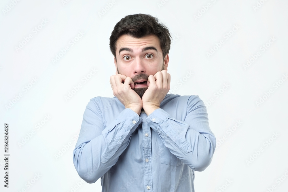 Young man dressed in casual clothes, biting nails and rounding eyes Young man dressed in casual clothes, biting nails and rounding eyes