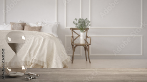 Wooden table, desk or shelf with crystal hourglass measuring the passing time in a countdown and house keys over vintage classic bedroom with soft bed, architecture interior design