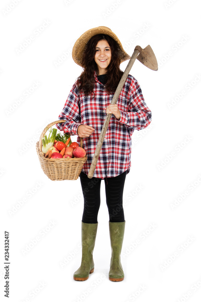 Young farmer with one arm holding a basket with fruit and vegetables, with the other a hoe, she is wearing a straw hat, checked shirt and green rubber boots