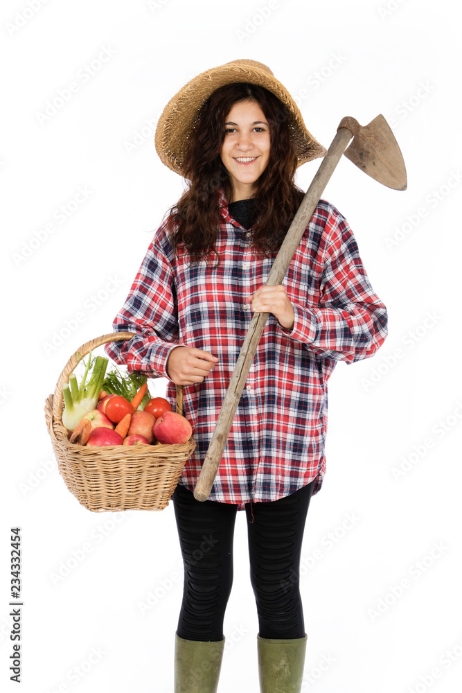 Young farmer with one arm holding a basket with fruit and vegetables, with the other a hoe, she is wearing a straw hat, checked shirt and green rubber boots