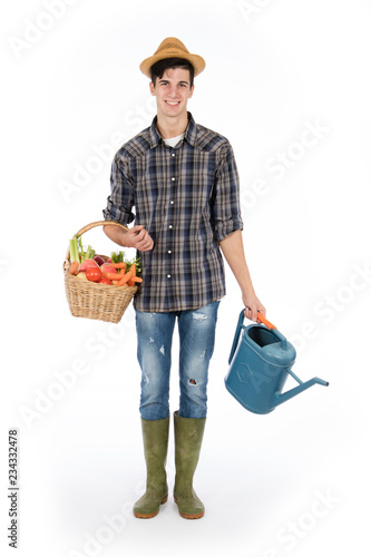 Young farmer with one arm holding a basket with fruit and vegetables, with the other a raincoat, he wears a straw hat, checked shirt and green rubber boots