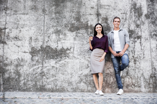 Calming couple man and woman, look at camera, make calm grimace, stand next to each other, have good relationship, on grey urban city street wall background. People, human facial expressions