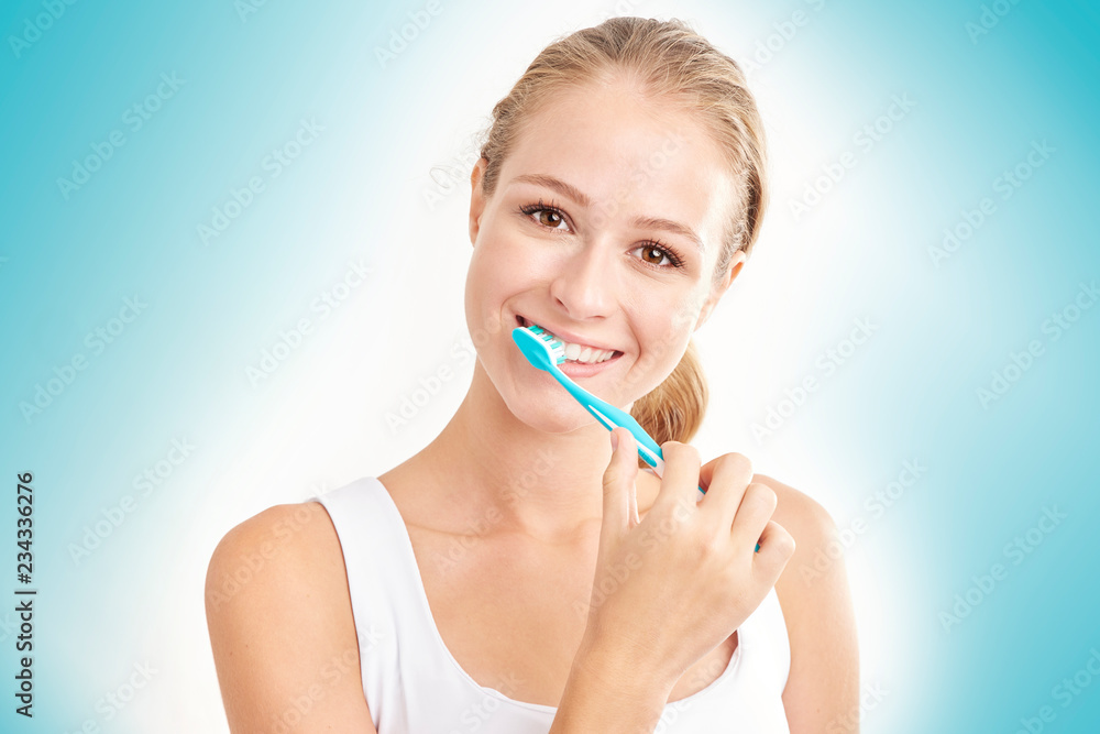 Studio shot of beautiful young woman brushing her teeth while looking at camera and smiling. Isolated on light blue background. Studio shot of beautiful young woman brushing her teeth while looking at camera and smiling. Isolated on light blue background.