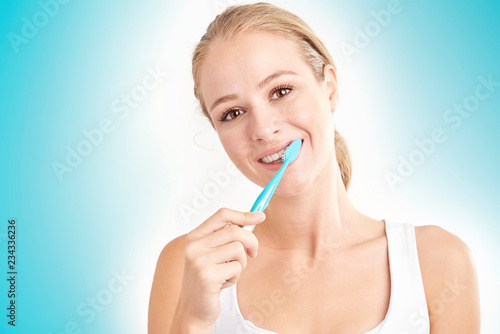 Studio shot of beautiful young woman brushing her teeth while looking at camera and smiling. Isolated on light blue background.